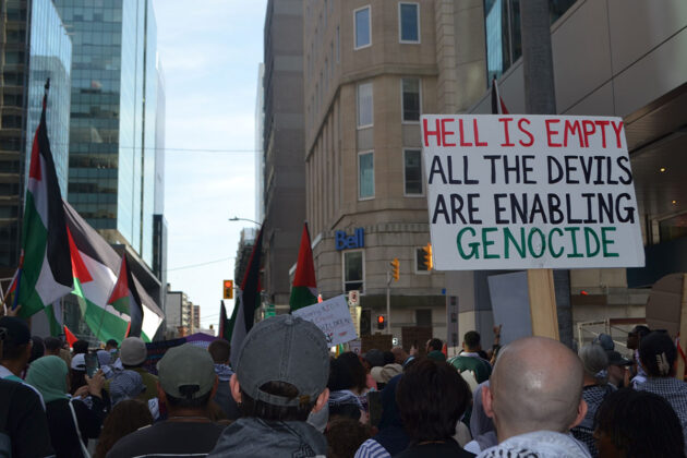 in a crowd of people, a person holds up a sign saying "hell is empty all the devils are enabling genocide"