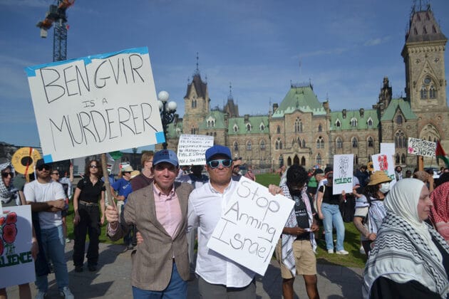 Two people stand in front of Parliament Buildings with signs saying "Bengvir is a murderer" and "Stop arming Israel"
