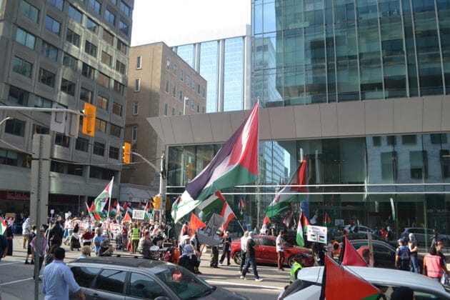 a crowd walks down a street with one person holding a large palestinian flag