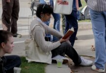In photos: With matcha, labubus and totes, Carleton crowns its most ‘performative male’ A man with headphones, a book and a totebag sits on the sidewalk curb.