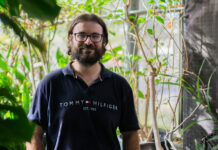 Q&A: Meet the new caretaker for thousands of Carleton’s butterflies A man in a dark blue Tommy Hilfiger shirt stands and smiles in a greenhouse.