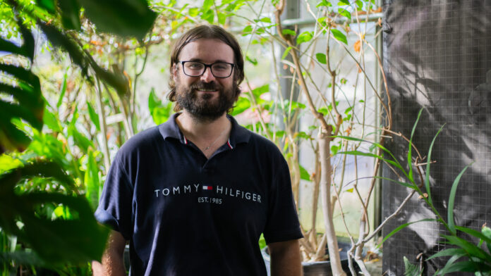 A man in a dark blue Tommy Hilfiger shirt stands and smiles in a greenhouse.