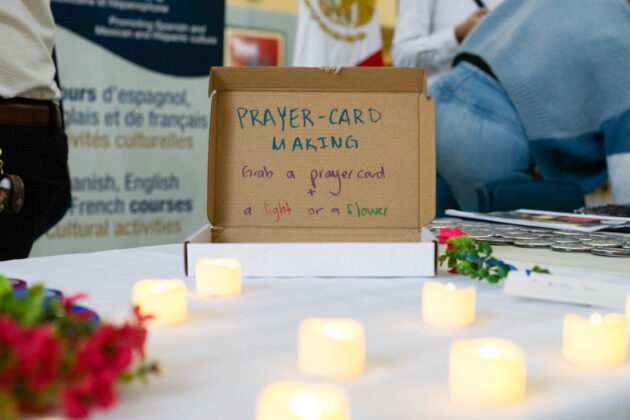 A cardboard box with a message written in marker on it saying "prayer-card making. Grab a prayer card + a light or a flower." Candles are in front of the box on a table.