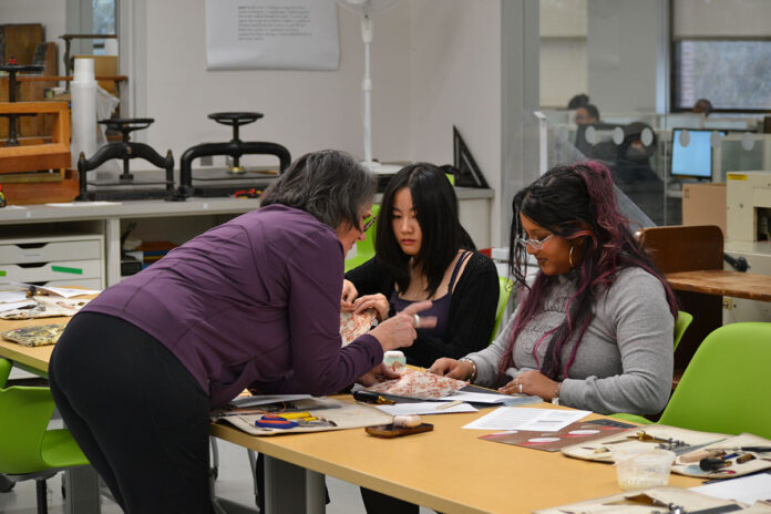 A woman leans over a table to talk to two younger people who are sitting with paper, scissors, thread and other tools in front of them on the table.