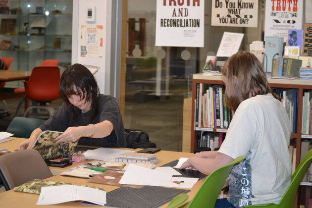 Two people sit at a table, one opening a book, the other hands on a paper.