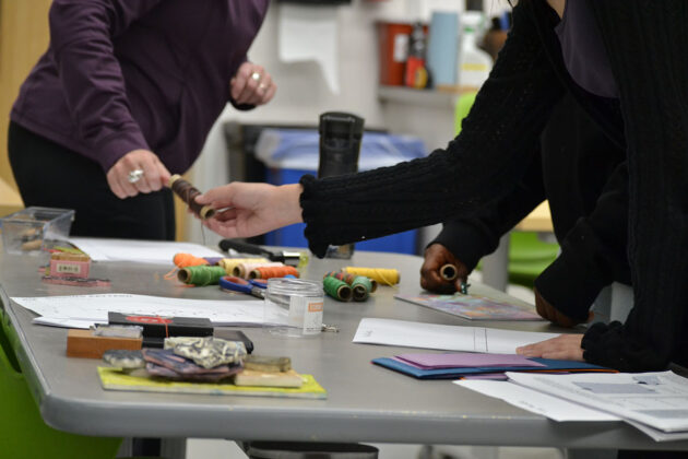 A hand passes a spool of brown thread to another hand over a table that has many spools of different-coloured thread, as well as jars, papers and scissors.