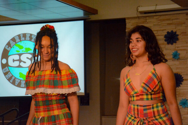 Two people stand on a stage, one in a dress, one in a two-piece outfit with a skirt and top. The outfits have a checkered pattern