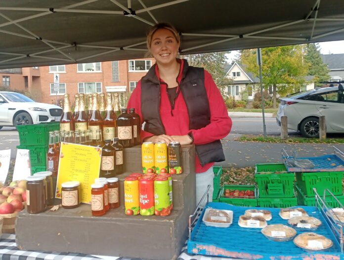 A woman standing in a red sweater and black jacket behind a stack of bottles.