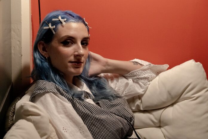 A person with blue hair sits and looks at the camera against an orange-painted wall.
