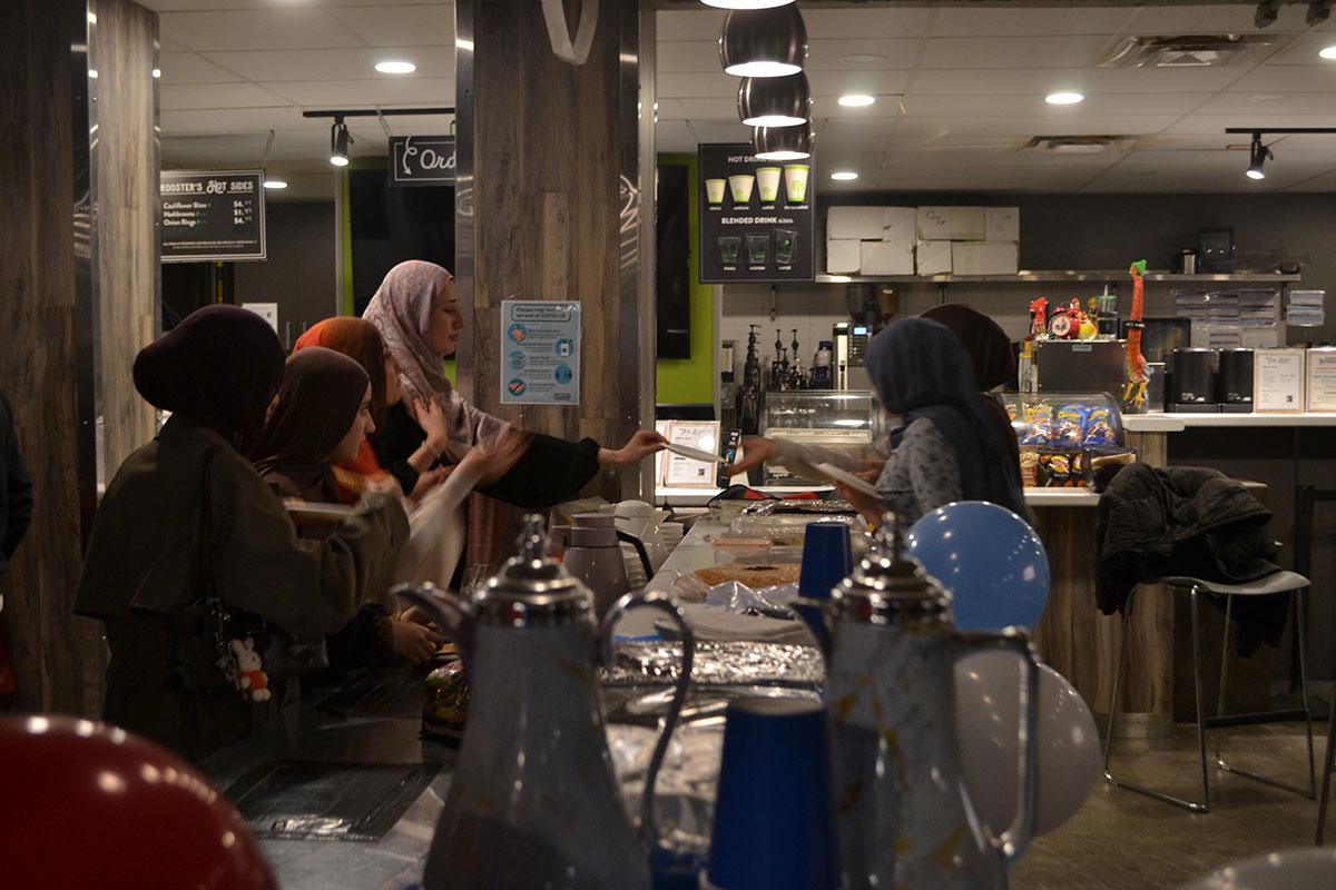 Several people stand on either side of a counter that has plates with food on them and teapots. One person passes a plate to another.