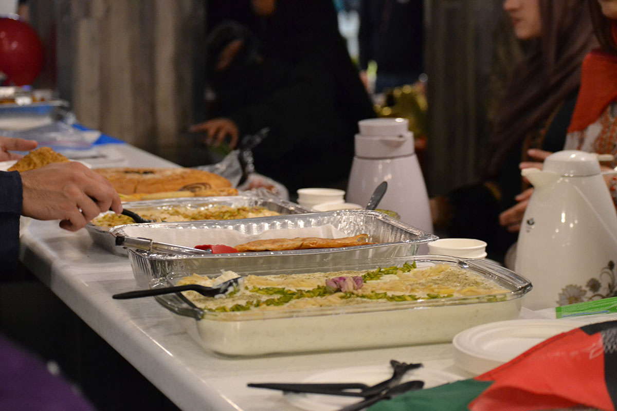 Platters and trays of food are on a counter filled with plastic cutlery and some kettles.