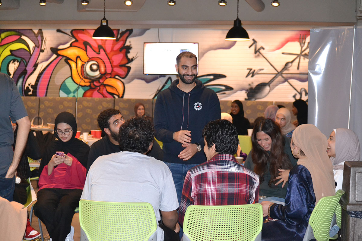 A group of people sit at a table, many laughing, with one person standing in the middle of the group.