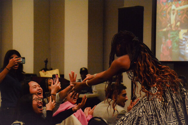 A person standing in a dress reaches her hands out to people reaching their hands out to her as she walks down a runway