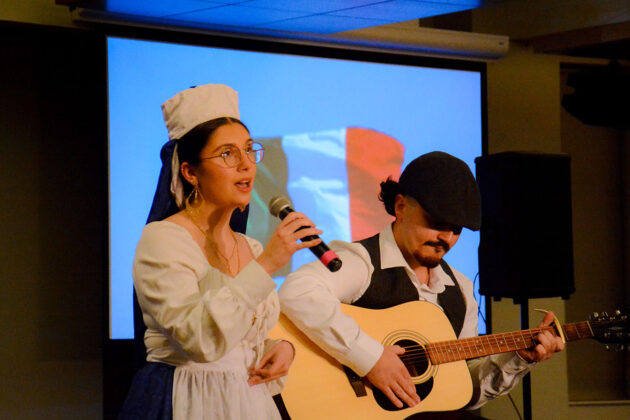 A person sits playing a guitar while another holds a microphone up to their mouth
