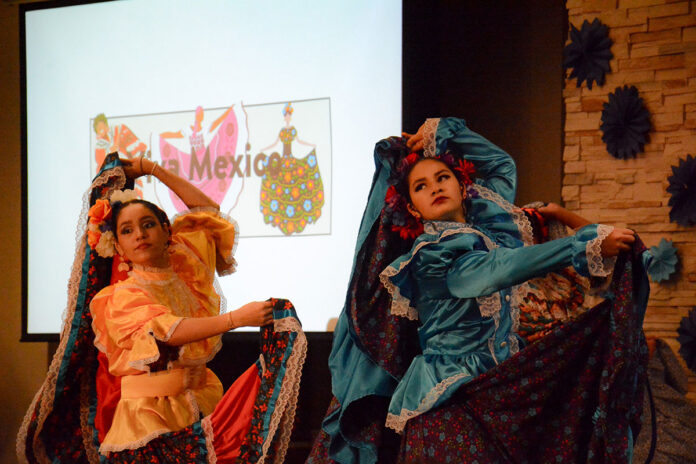 Two people hold up their long skirts on their dresses up above their heads as they dance on a stage