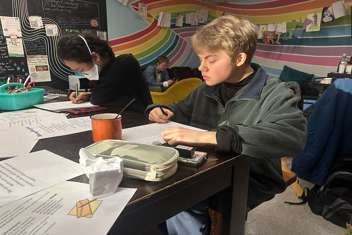 A person sits at a table and writes on a piece of paper. Somebody next to them does the same. In the background, the wall has a rainbow design, and there are several papers hanging