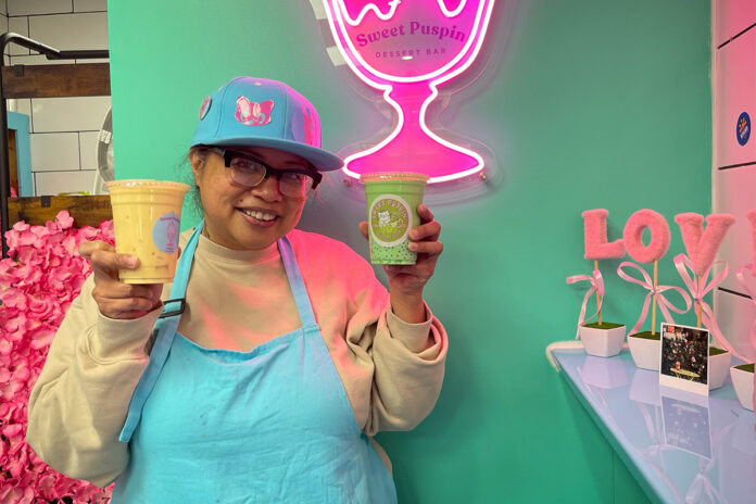 A woman wearing an apron stands and holds two cups with liquids in them in front of a light-up decoration of a cup that says 