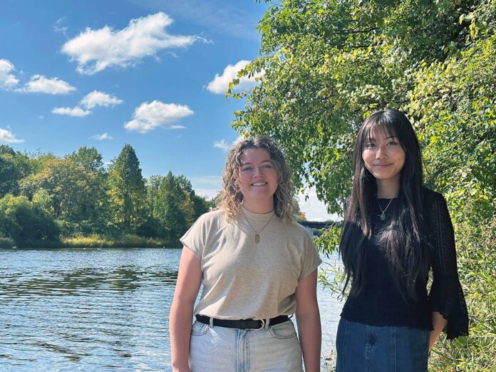 Two women stand in front of a clearing near the Carleton University campus smiling.