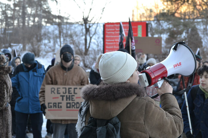 A person holds a megaphone to their mouth in front of a crowd of people, some holding signs, including one that says 