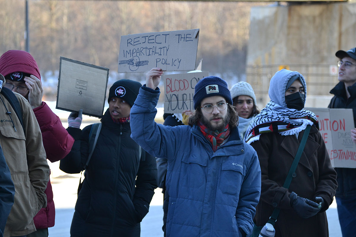 People stand wearing jackets and tuques, many holding signs including one that says "reject the impartiality policy"