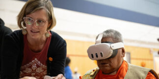 ‘Keep the fire burning’: How holograms and AI are helping strengthen this First Nation’s language and culture An elderly man sits with a virtual reality mask covering his eyes. He holds a remote that a woman standing next to him is also controlling with her hand.