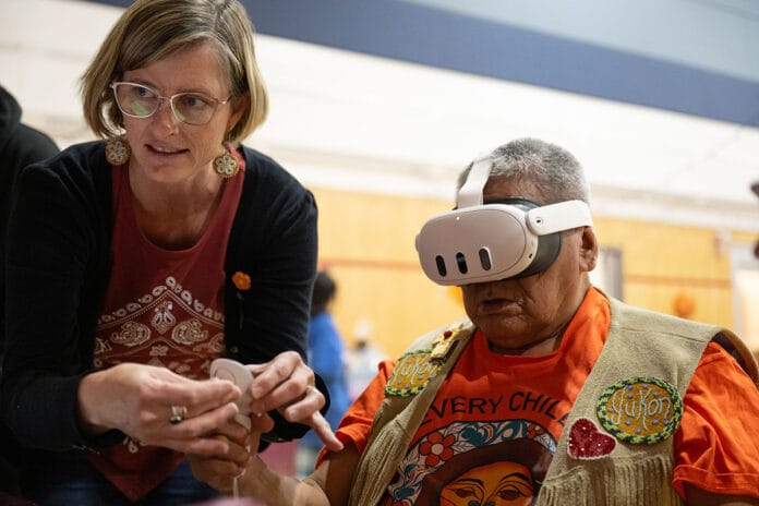 An elderly man sits with a virtual reality mask covering his eyes. He holds a remote that a woman standing next to him is also controlling with her hand.