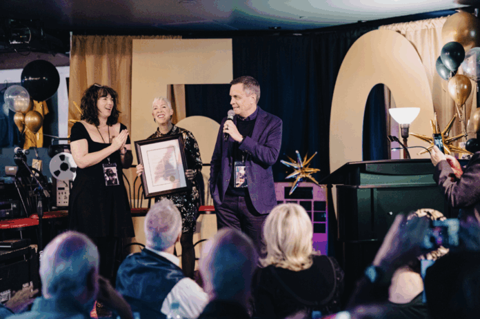 An audience with a man speaking and two woman by his side