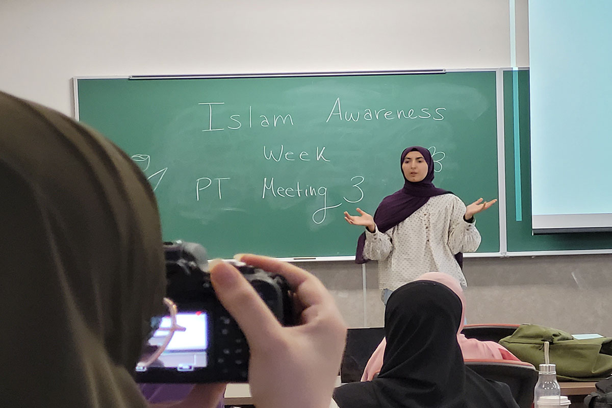 A woman stands in front of a chalkboard that has "Islam Awareness Week PT Meeting 3" written on it.