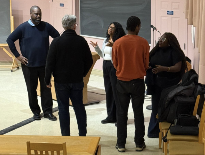 Five people - three facing to the camera and two facing away - are engaging in a seemingly vivacious conversation in a university classroom. They are all standing.