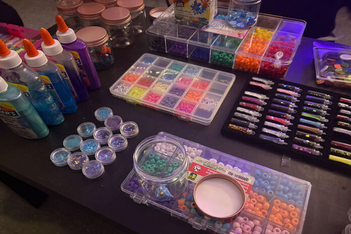 Glitter glue, glitter and beads and buttons in a variety of shapes, sizes and colours sit on a table in a low-lit room