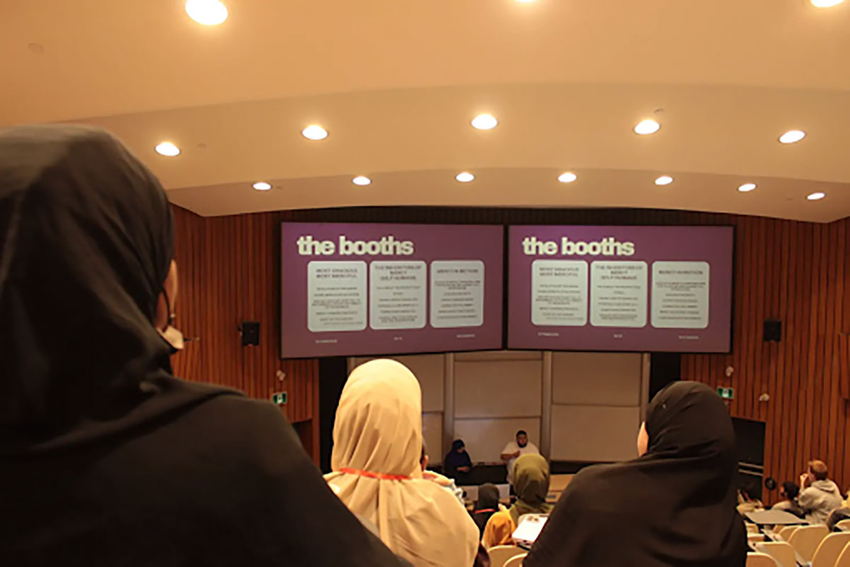 A crowd of people watch presenters at the front of the room below a large screen that has the title "the booths"
