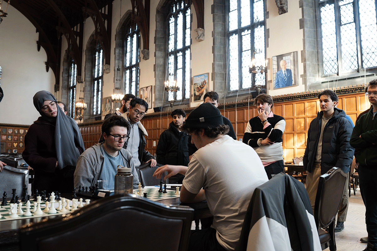 A small crowd, including one woman stand around a table where two men are playing chess.