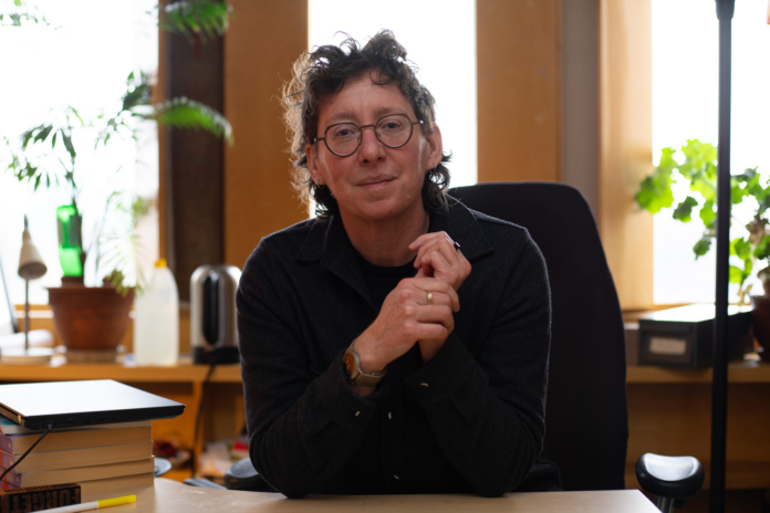 A person sits in an office with a stack of books on their desk and plants in the background.