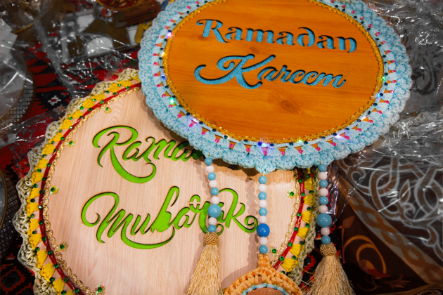 Two wooden signs sit on a table. One sign says "Ramadan Mubarak" and the other says "Ramadan Kareem." There are beads and different decorations on the circular signs.
