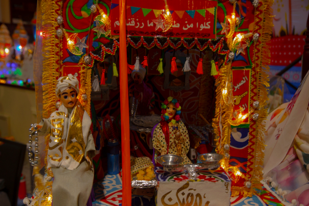 Two dolls, one man and one woman, sit in a mini booth with bright curtains and decorations.