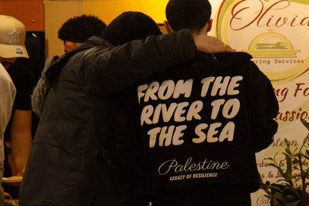Two men have their backs to the camera. One has an arm over the others' shoulder, who's wearing a sweater that reads "From the river to the sea, Palestine legacy of resilience."