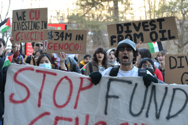 A crowd stands together outside, many holding signs, including ones that say "We CU investing in genocide," "$34M in genocide," and "Divest from oil."