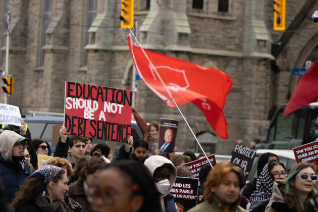 A crowd of people hold signs and a flag waves in the air. One sign says "Education should not be a debt sentence"