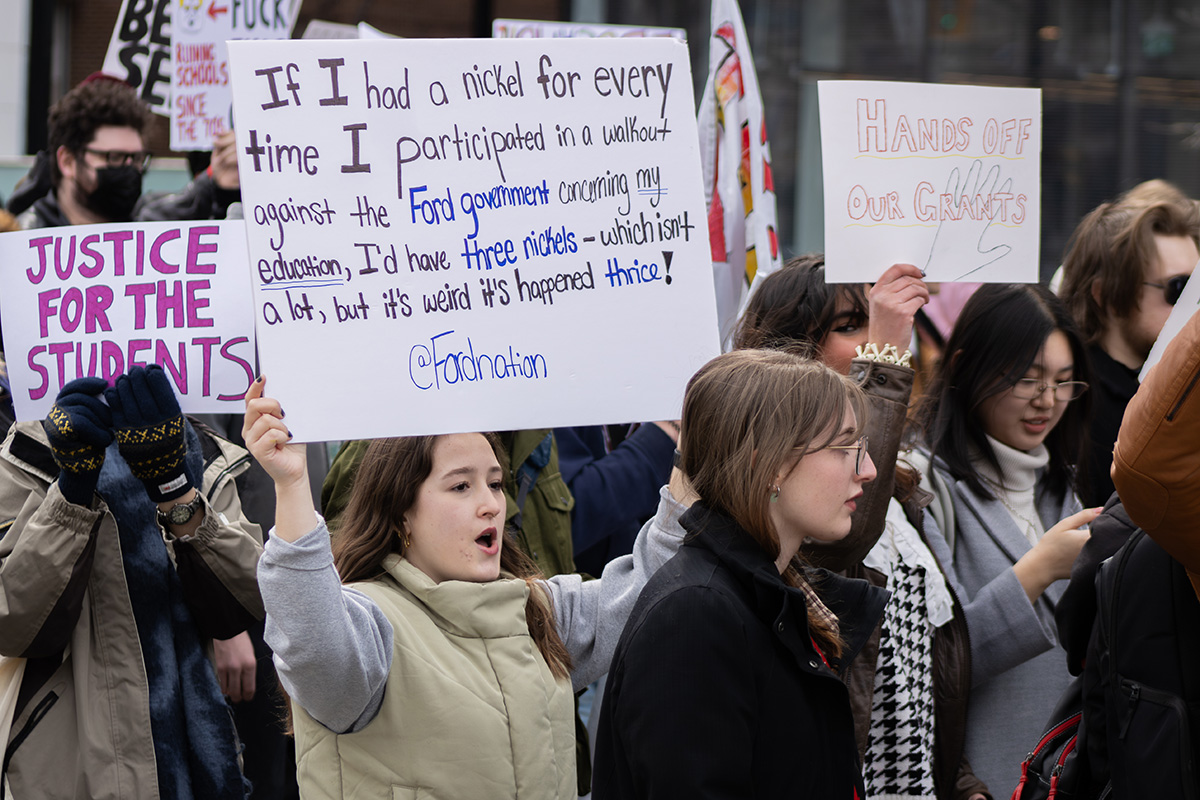 A woman stands in a crowd where she holds a sign that says "If I had a nickel for every time I participated in a walkout against the Ford government concerning my education, I'd have three nickels - which isn't a lot, but it's weird it's happened thrice! @Fordnation"
