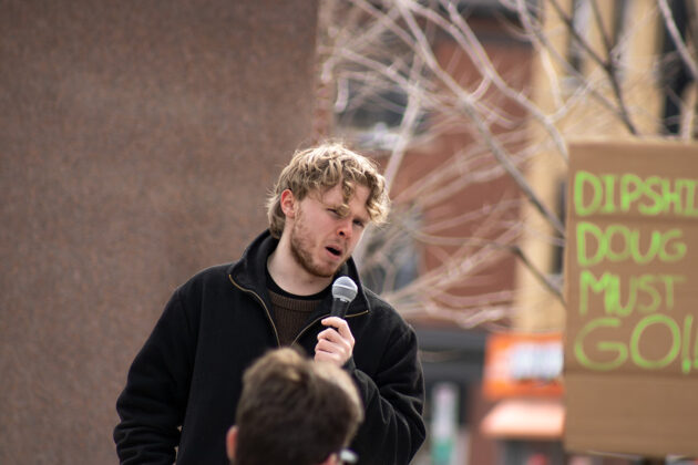 A man holds a microphone up to his mouth. Behind him, there is a sign that says "Doug must go!"