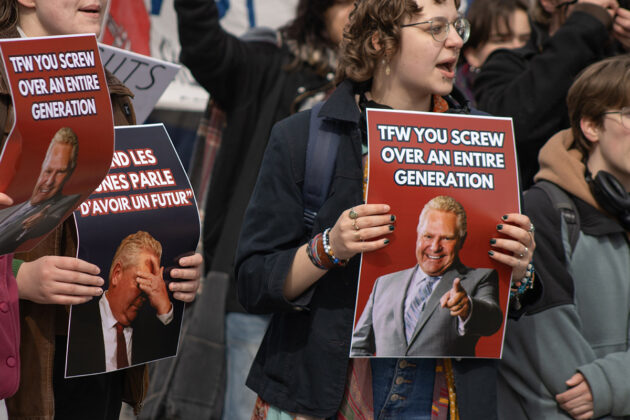 A person stands in a crowd holding a sign that has a photo of Doug Ford pointing and smiling with the words "TFW you screw over an entire generation"