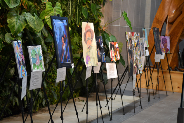 Eleven paintings/drawings sit on stands in a row. Most of the art pieces show people on them.