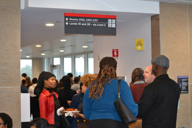 Dozens of people line up in a hall, some holding plates of food and speaking to each other