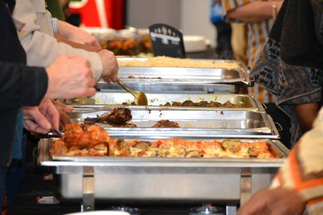 Trays of lasagna, chicken, curried goat sit on a table as people serve themselves with spoons.