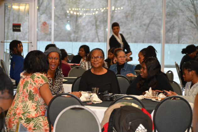 Groups of people sit at tables with plates of food in front of them. Some laugh, some talk to each other.