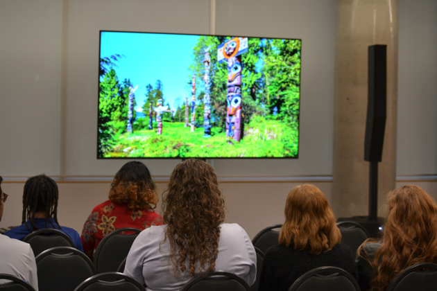 The backs of people's heads are visible as they face a screen that has an outdoor scene with totem poles standing