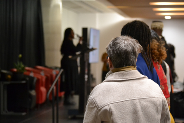 A person stands at a microphone on stage as spectators stand