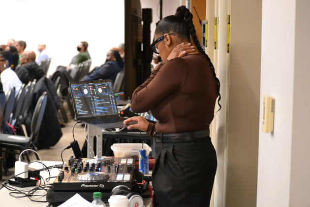 A woman stands in front of a laptop and soundboard at the back of a crowd of people sitting