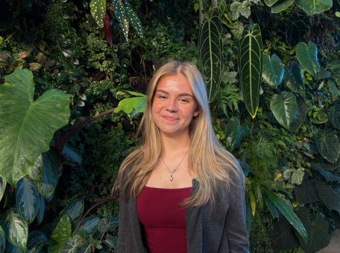 A woman is standing in front of the green plant wall.
