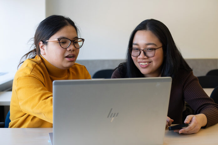 Two women sit next to each other behind a table, looking at a laptop screen. The woman on the left points at a laptop screen.
